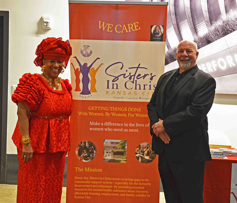Carolyn Whitney with a man standing in front of a banner of Sisters in Christ and the info about the organisation